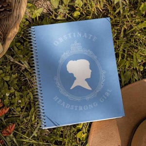 May include: A light blue spiral-bound notebook with the words "OBSTINATE HEADSTRONG GIRL" and a woman's profile silhouette. The notebook is on green grass.