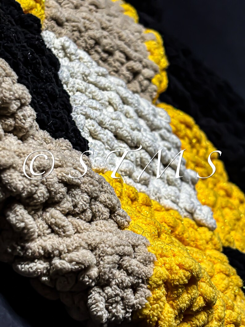 May include: A close-up of a crocheted flower made with yellow, white, black and brown yarn. The flower has a textured surface and is made with a variety of stitches.