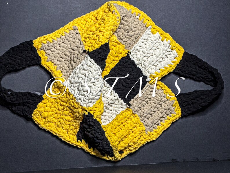 May include: A crocheted tote bag made with black, yellow, white and beige yarn. The bag features a diamond pattern and two black straps.