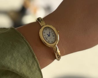 May include: A gold-tone bangle watch featuring an oval-shaped case with a white dial, black Roman numeral hour markers, and blue hands. The watch has a smooth, polished metal band that encircles the wrist, providing a sleek and simple appearance.