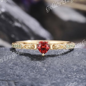May include: A gold ring featuring a central heart-shaped red garnet gemstone. The band displays an intricate filigree design on both sides of the stone, accented with small, light green round-cut gemstones embedded along the shank of the ring.
