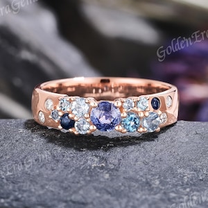 May include: Rose gold ring featuring a band of blue gemstones, with a central purple stone. The ring is displayed on a dark grey stone surface. The text "Golden Era Gems" is visible.