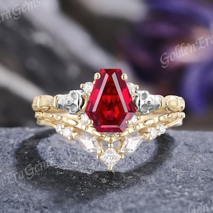 May include: A gold-toned ring set featuring a large, faceted, red gemstone in a coffin shape. The band is adorned with small skulls and white gemstones. The set includes a matching band with a similar design. The text "Golden Era Gems" is visible.