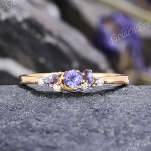Peut inclure: Une bague en or ornée d'une pierre précieuse violette ronde au centre, flanquée de pierres violettes et blanches plus petites. La bague est posée sur une surface en pierre gris foncé, avec le texte "Golden Era Gems".
