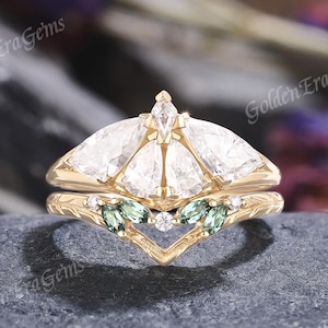 May include: A gold engagement ring set with a unique design. The ring features a cluster of pear-shaped clear stones and smaller green stones. The band has a delicate, engraved pattern. The ring is set on a dark gray surface.
