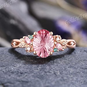 May include: Rose gold ring featuring a large, oval-cut pink gemstone. The ring is adorned with leaf and floral accents, small teal stones, and tiny white gems. The words "Golden Era Gems" are printed on the image.