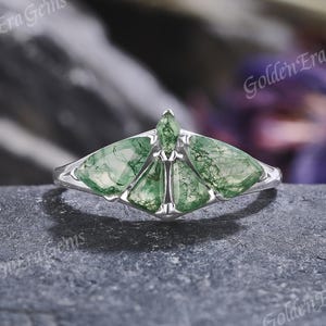May include: A silver ring featuring a fan-shaped arrangement of five moss agate stones with green inclusions. The stones are set in a polished metal band, creating a symmetrical design that resembles a moth or butterfly shape with clear green patterns.