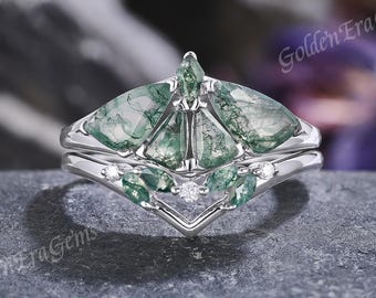 Op de afbeelding: Een zilveren ringenset met groene mosagaatstenen en kleine, heldere accentstenen. De ring heeft een kenmerkende V-vormige band. De tekst "Golden Era Gems" is zichtbaar. De set staat tegen een vage achtergrond.