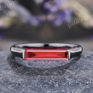 May include: A silver ring with a rectangular, vibrant red gemstone. The ring's band is smooth and polished, and the gemstone is set horizontally. The ring is displayed on a textured gray surface. The text "Golden Era Gems" is visible.