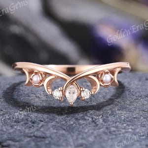 May include: Rose gold ring with a curved design. The ring features a pear-shaped center stone, two smaller round stones, and two pearls. The ring is set on a dark gray surface.