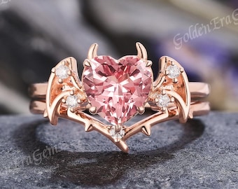 May include: Rose gold engagement ring with a heart-shaped pink gemstone. The ring features small diamond accents and a unique design with horns and wings. The ring is set on a dark gray stone surface.