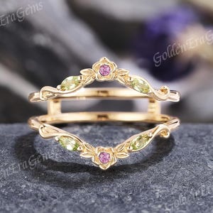 May include: A gold-colored ring guard with a floral and leaf design. The ring features small, oval-shaped green gemstones and pink flower accents. The ring is set against a dark gray stone surface.