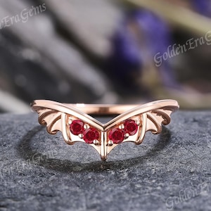 May include: Rose gold ring with a wing design and three red gemstones. The ring is on a gray background.
