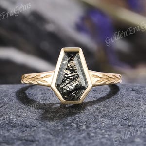 May include: A gold ring with a unique coffin-shaped gemstone. The gemstone is a clear, faceted stone with black inclusions. The ring band features a delicate leaf design on the shoulders. The ring is set on a dark gray stone surface.