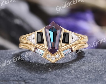 May include: A gold ring featuring a central kite-shaped color-changing gemstone flanked by two black baguette stones and two small triangular stones. The band includes additional small clear stones and text reading Golden'EraGems.