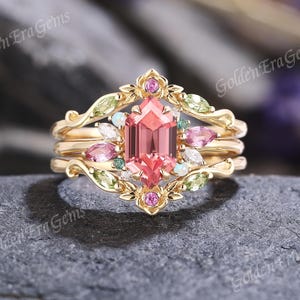 May include: A three-piece gold ring set. The center ring features a large, pink, hexagonal gemstone. The set is adorned with smaller pink, green, and white gemstones. The rings have a floral and leaf design.