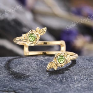 May include: Two gold rings with a floral design. Each ring features a green gemstone center surrounded by gold petals and leaves. The rings are displayed on a dark gray, textured surface.