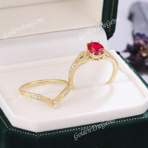 Könnte beinhalten: Ein Goldring-Set mit einem zentralen, oval geschliffenen roten Edelstein in einer Krappenfassung auf einer Ringschiene mit filigranen Details, ergänzt durch einen passenden V-förmigen Chevron-Ring mit kleinen klaren Steinen an der Oberseite.