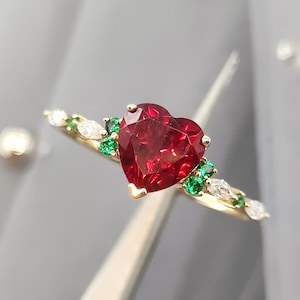 Puede incluir: Un anillo de oro con un rubí en forma de corazón y piedras preciosas verdes y blancas.