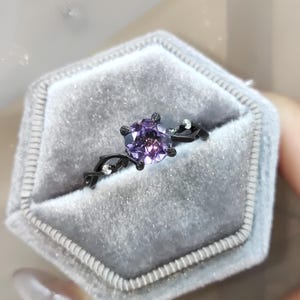 May include: A black ring with a purple gemstone set in a delicate, vine-like design. The ring is displayed in a gray velvet ring box.