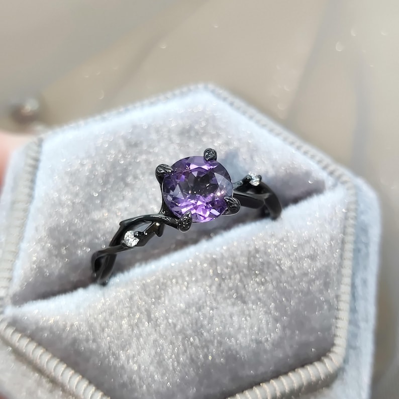 May include: A black ring with a purple gemstone set in a twisted vine design. The ring is in a black velvet ring box.