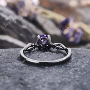 May include: Black engagement ring with a purple amethyst gemstone and intricate detailing. The ring is set on a black background.