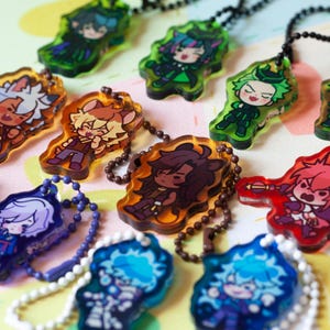 Peut inclure: Une collection de porte-clés en acrylique translucide représentant divers personnages d'anime de style chibi dans des couleurs vives, notamment rouge, bleu, vert et ambre. Chaque porte-clés est fixé à une petite chaînette à billes en métal.