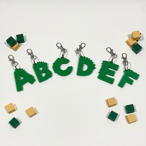 Puede incluir: Seis abalorios de letras verdes con un borde de hojas, cada uno en un llavero plateado. Las letras deletrean "ABCDEF".