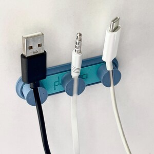 Puede incluir: Un organizador de cables azul con tres ranuras, que sostiene un cable USB negro, un cable jack de audio blanco y un cable USB-C blanco. El organizador tiene la palabra "pl" impresa.