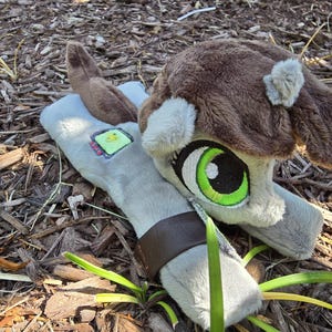 Puede incluir: Un peluche marrón y gris con grandes ojos verdes y una gorra marrón. El juguete tiene un brazo gris claro con una correa marrón y un pequeño parche con un cuadrado verde. Está sobre un lecho de virutas de madera.