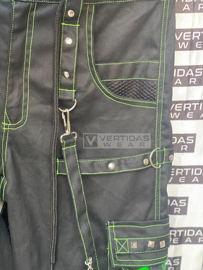 May include: Black pants with green stitching and a mesh pocket. The pants have a zipper fly and a belt loop. The pants have a large pocket on the right leg with silver studs.