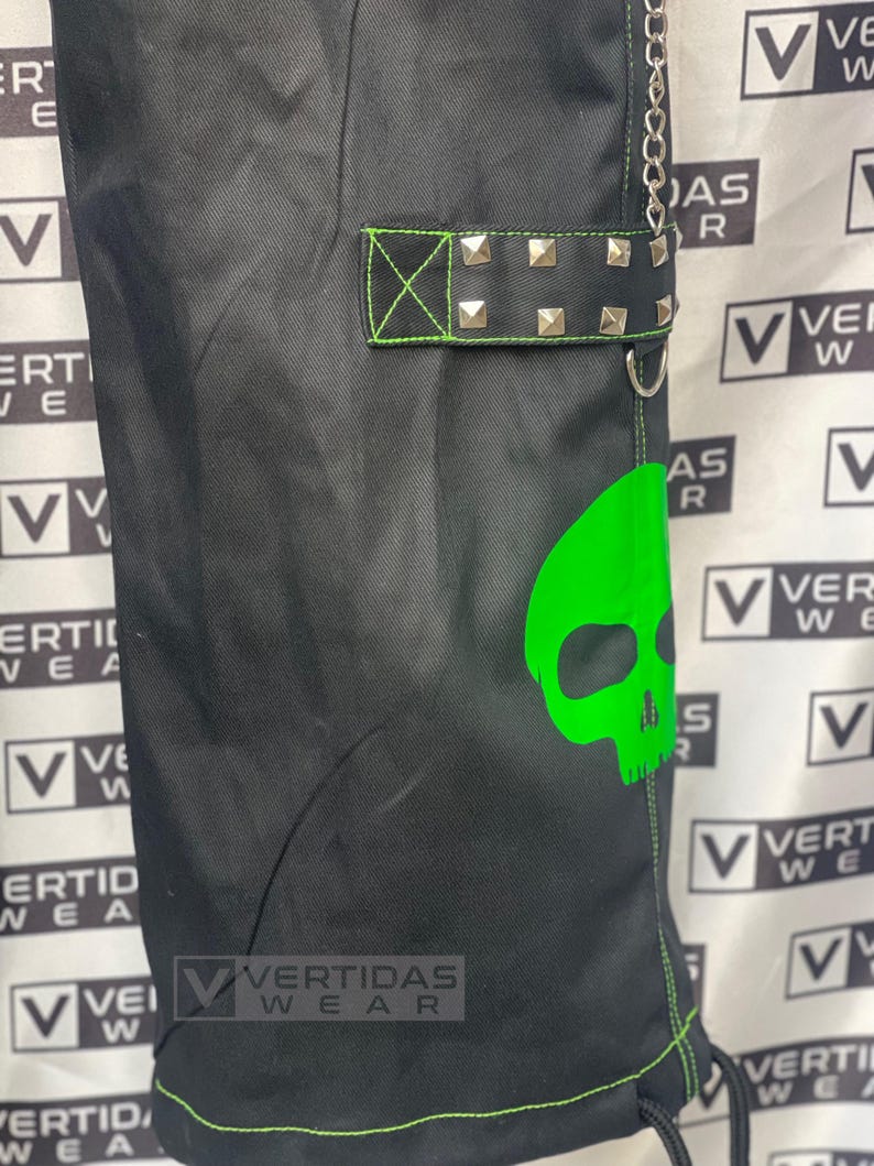May include: Black cargo pants with a green skull graphic and a black belt with silver studs. The pants have a green drawstring and the text "VERTIDAS WEAR" is visible on the pants.