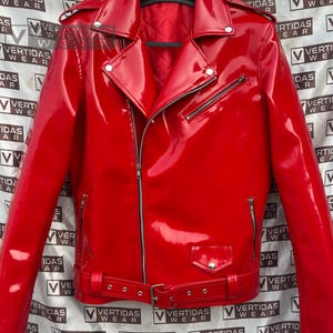 Puede incluir: Chaqueta de moto roja brillante con cierre de cremallera asimétrico clásico, cuello con solapas y cintura con cinturón. La chaqueta presenta cremalleras y herrajes plateados, y un pequeño bolsillo. La chaqueta está colgada de una percha negra.