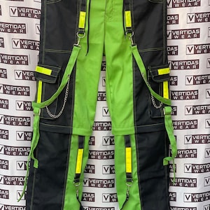 Könnte beinhalten: Schwarze und neongrüne Cargohose mit Hosenträgern. Die Hose hat ein zweifarbiges Design, mit Schwarz an den Seiten und Neongrün in der Mitte. Gelbe reflektierende Streifen und silberne Kettendetails setzen Akzente. Mehrere Taschen.