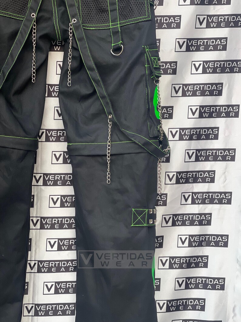 May include: Black cargo pants with green stitching and silver chains. The pants have multiple pockets and a mesh panel on the front. The pants are labeled "Vertidas Wear" on the side.