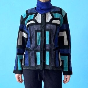 May include: A geometric patterned jacket in shades of blue, teal, gray, and black, paired with black pants and a blue turtleneck. The jacket features a button-up front and a unique patchwork design. The outfit is completed with blue shoes.