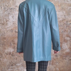 May include: A teal-colored leather jacket with a V-shaped design on the back. The jacket has long sleeves and a collared neckline. The person is wearing black and white checkered pants.