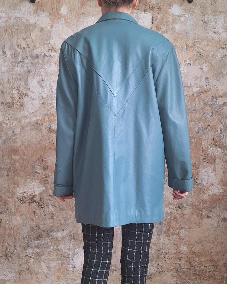 May include: A teal-colored leather jacket with a V-shaped design on the back. The jacket has long sleeves and a collared neckline. The person is wearing black and white checkered pants.