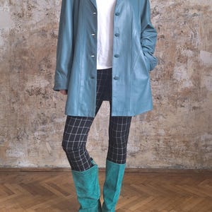 May include: A light blue leather jacket with a button-up front, worn over a white t-shirt and black and white plaid leggings. The outfit is completed with turquoise suede knee-high boots. The background is a textured wall.