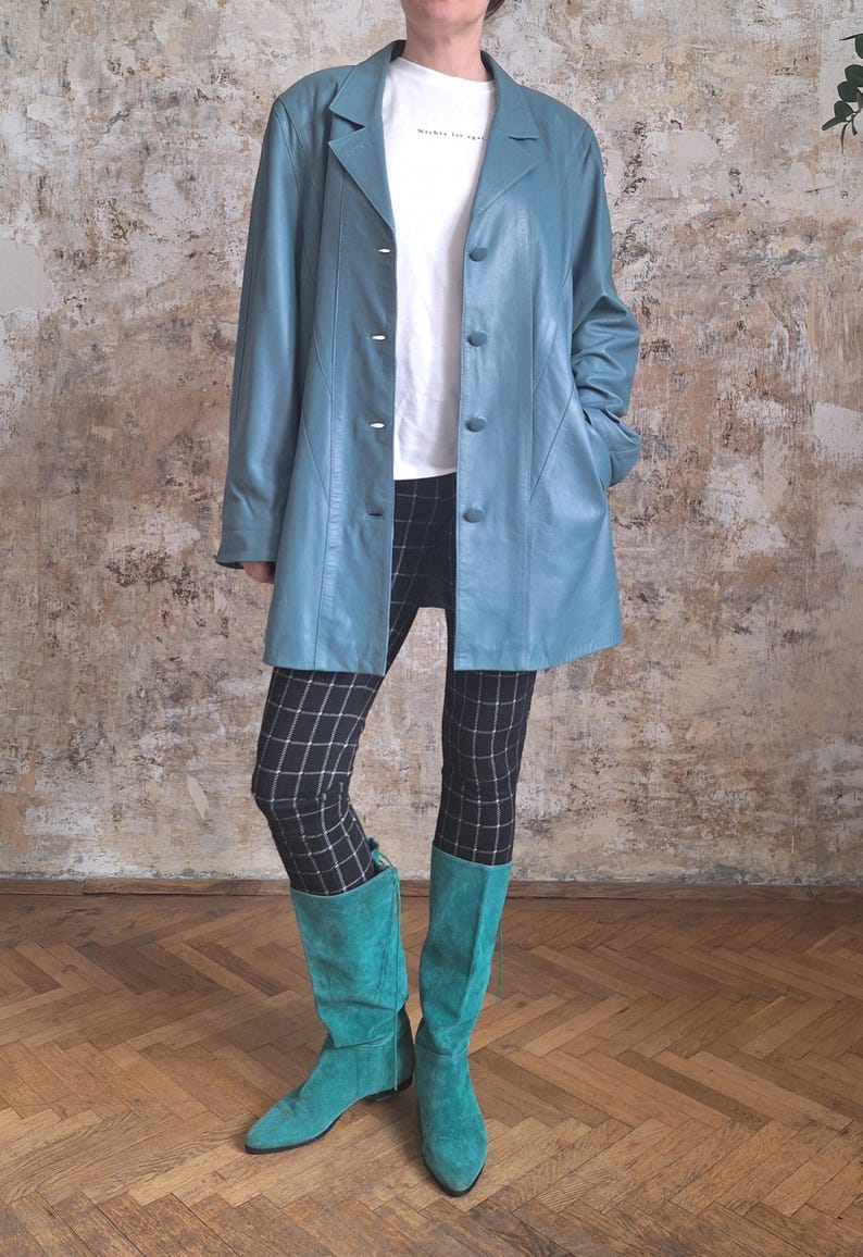 May include: A light blue leather jacket with a button-up front, worn over a white t-shirt and black and white plaid leggings. The outfit is completed with turquoise suede knee-high boots. The background is a textured wall.