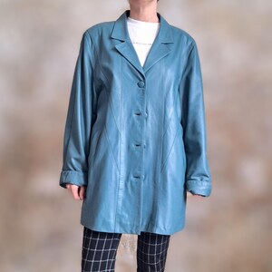 May include: A teal leather coat with a classic lapel collar and three-button closure. The long-sleeved coat has a tailored fit and a knee-length hemline. The coat is worn over a white t-shirt and black and white checkered pants.