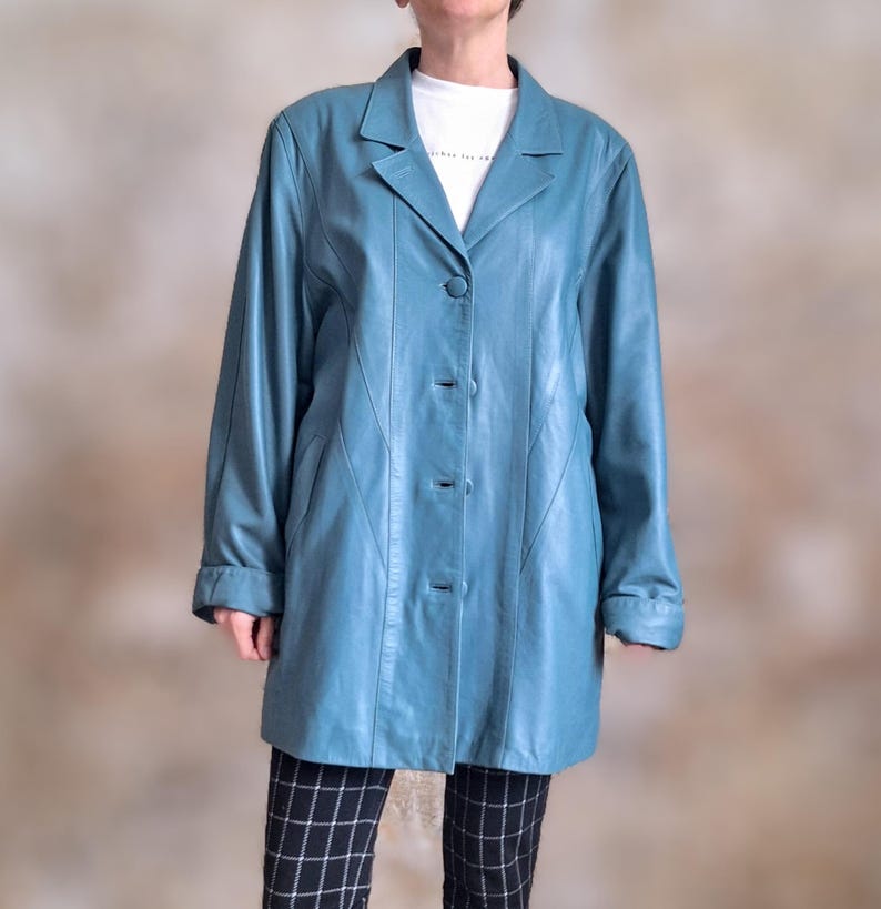 May include: A teal leather coat with a classic lapel collar and three-button closure. The long-sleeved coat has a tailored fit and a knee-length hemline. The coat is worn over a white t-shirt and black and white checkered pants.