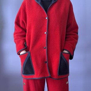 May include: A red fleece jacket and matching pants set. The jacket has black trim, pockets, and buttons. The pants have gold-colored buttons on the side. The person is wearing a black turtleneck. This outfit is suitable for cold weather.