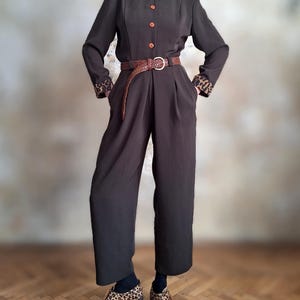 Könnte beinhalten: Dunkelbrauner Jumpsuit mit Leopardenmuster an Kragen und Manschetten. Der Jumpsuit hat einen braunen Ledergürtel mit goldener Schnalle. Die Schuhe sind im Leopardenmuster. Vintage-Stil.