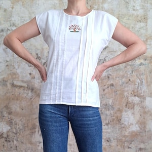 Pode incluir: Top de linho branco com gola redonda e mangas curtas. A parte superior apresenta pregas verticais na frente e um pequeno desenho de árvore bordado. A modelo está usando jeans azuis.