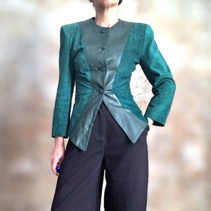 May include: A teal green suede and leather jacket with a unique color-blocked design. The jacket features a round collar, long sleeves, and three buttons down the front. The model is wearing black pants.