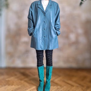 May include: A light blue leather coat with a classic collar and button closure. The coat is paired with black and white checkered pants and turquoise knee-high boots. The outfit is set against a neutral background and wooden floor.