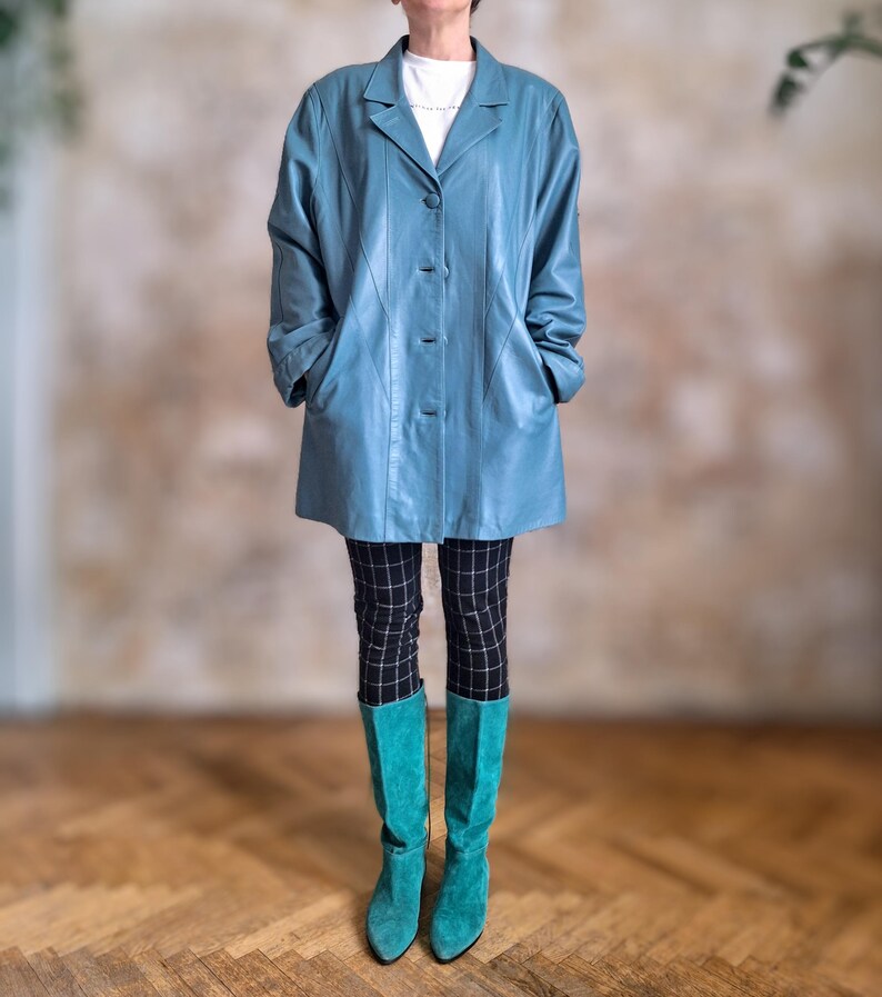May include: A light blue leather coat with a classic collar and button closure. The coat is paired with black and white checkered pants and turquoise knee-high boots. The outfit is set against a neutral background and wooden floor.