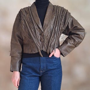 May include: A brown leather jacket with a unique pleated design on the shoulders and upper chest. The jacket has a notched collar, two buttons, and long sleeves. The person is wearing a black turtleneck and blue jeans.