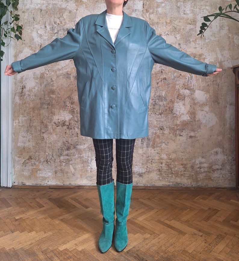 May include: A teal-colored leather coat with a button-up front and a classic collar. The coat is paired with black and white checkered pants and teal boots. The outfit is displayed on a wooden floor.